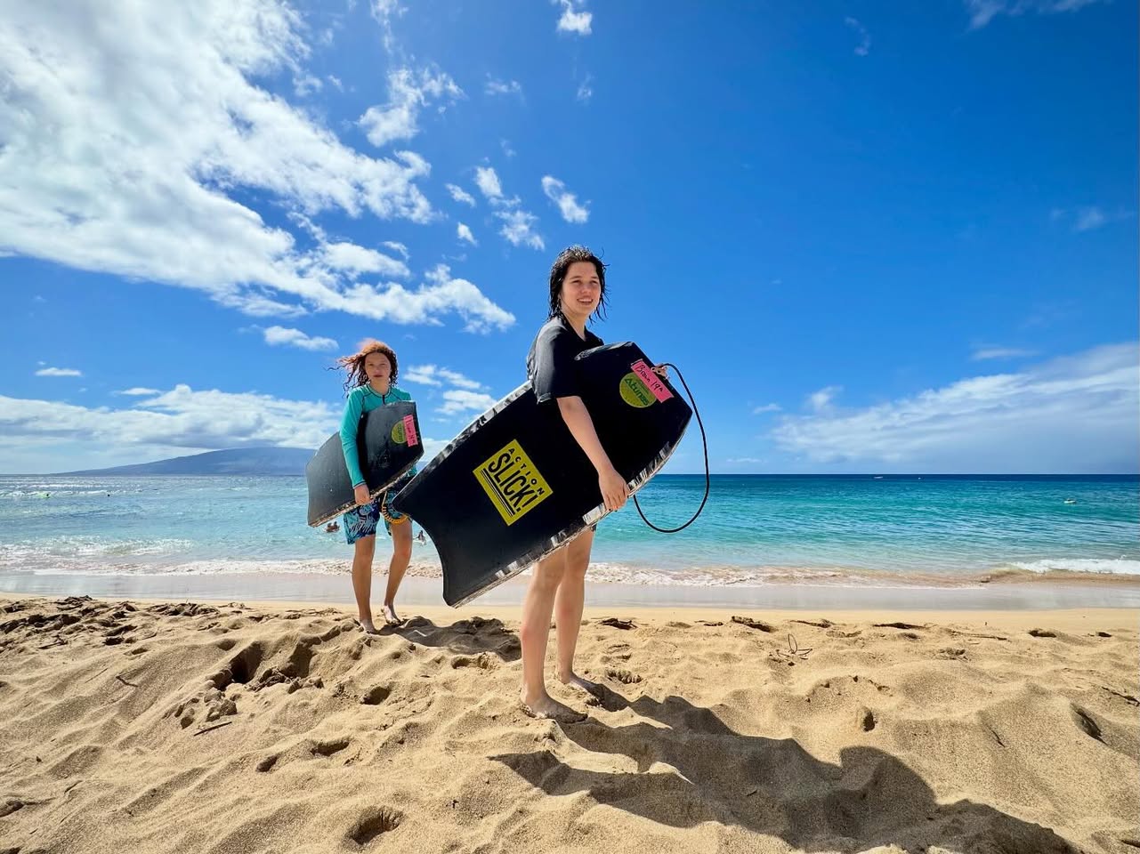 Two people holding paddleboards on a sandy beach under a bright blue sky.