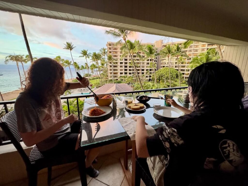Two people enjoying a meal on a balcony overlooking a tropical coastline with palm trees and a hotel building. A detailed view shows the scenic landscape and the dining setup.