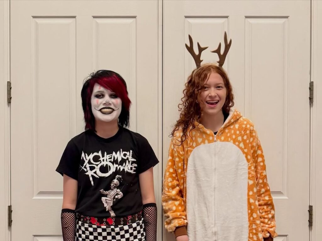 Two costumed family members standing together in front of a white interior door. One wears a punk rock outfit with face paint, and the other a reindeer onesie.