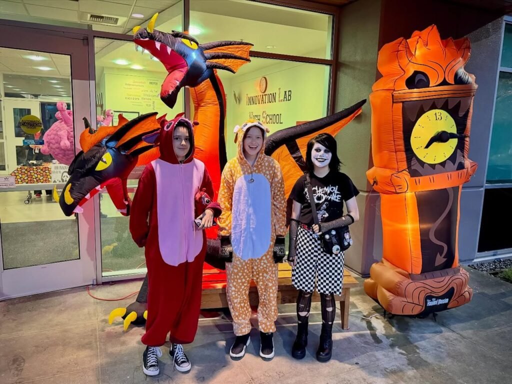 Three individuals in whimsical Halloween costumes pose with colorful inflatable decorations. One wears a full-body Spongebob look while others dress as a dragon and a character with a white mask.