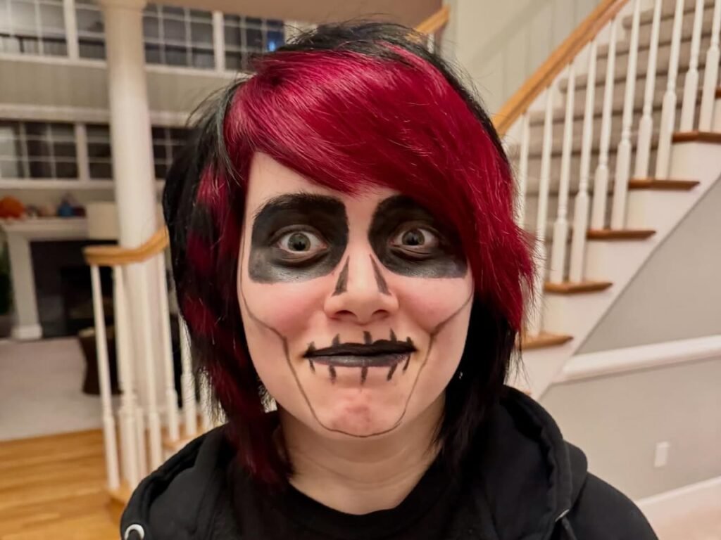 A person with red and black hair wears elaborate face paint resembling a Cheshire Cat with exaggerated eyes and a wide grin.