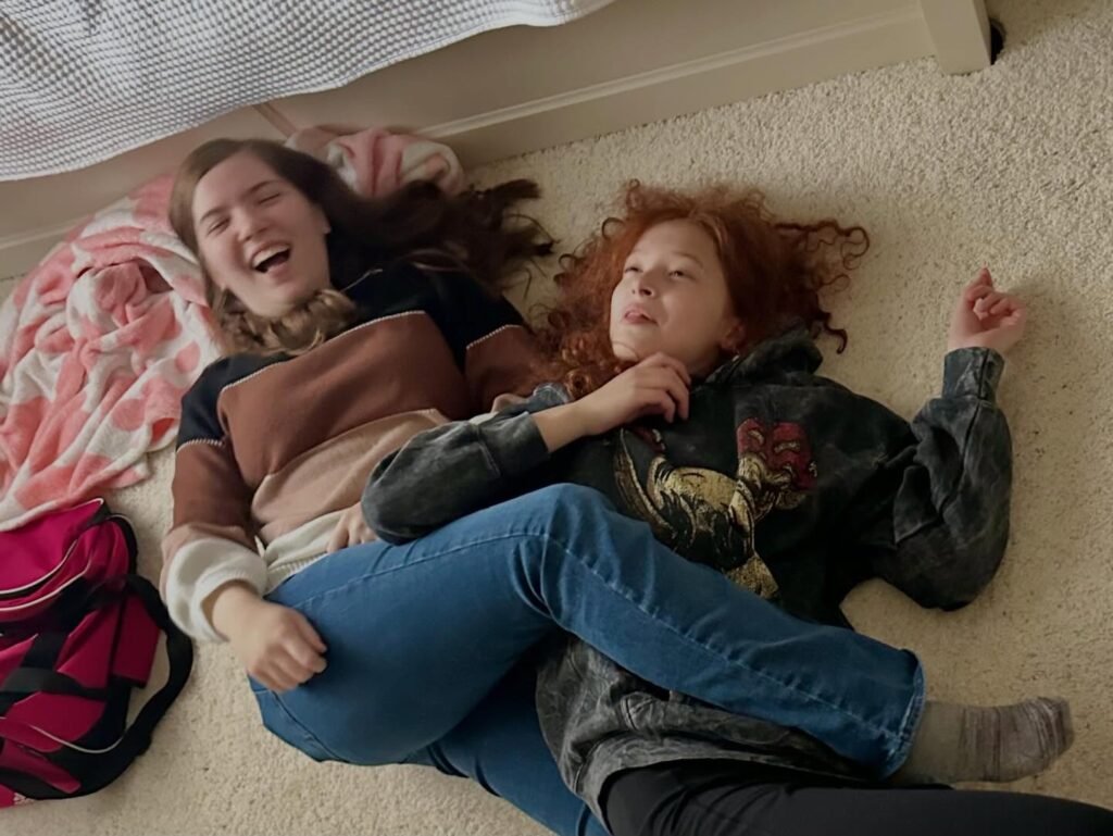 Two children lying on a carpeted floor playing together. A pink blanket and red backpack add color to the scene.