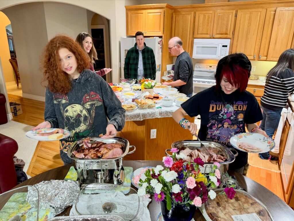 A group of people gather in a warm kitchen for a dinner party, serving food from chafing dishes. Individuals hold plates while others prepare in the background, surrounded by homey wooden cabinets.