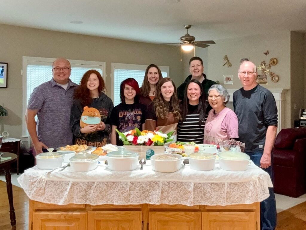 A group of smiling friends and family members pose behind a buffet table laden with holiday dishes and a colorful bouquet.