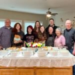 A group of smiling friends and family members pose behind a buffet table laden with holiday dishes and a colorful bouquet.