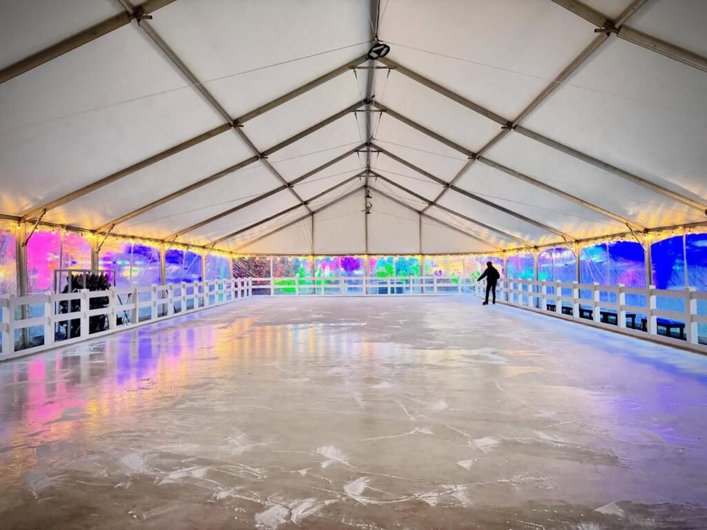 A spacious indoor ice skating rink with vibrant neon lights glowing along the walls and ceiling structure.