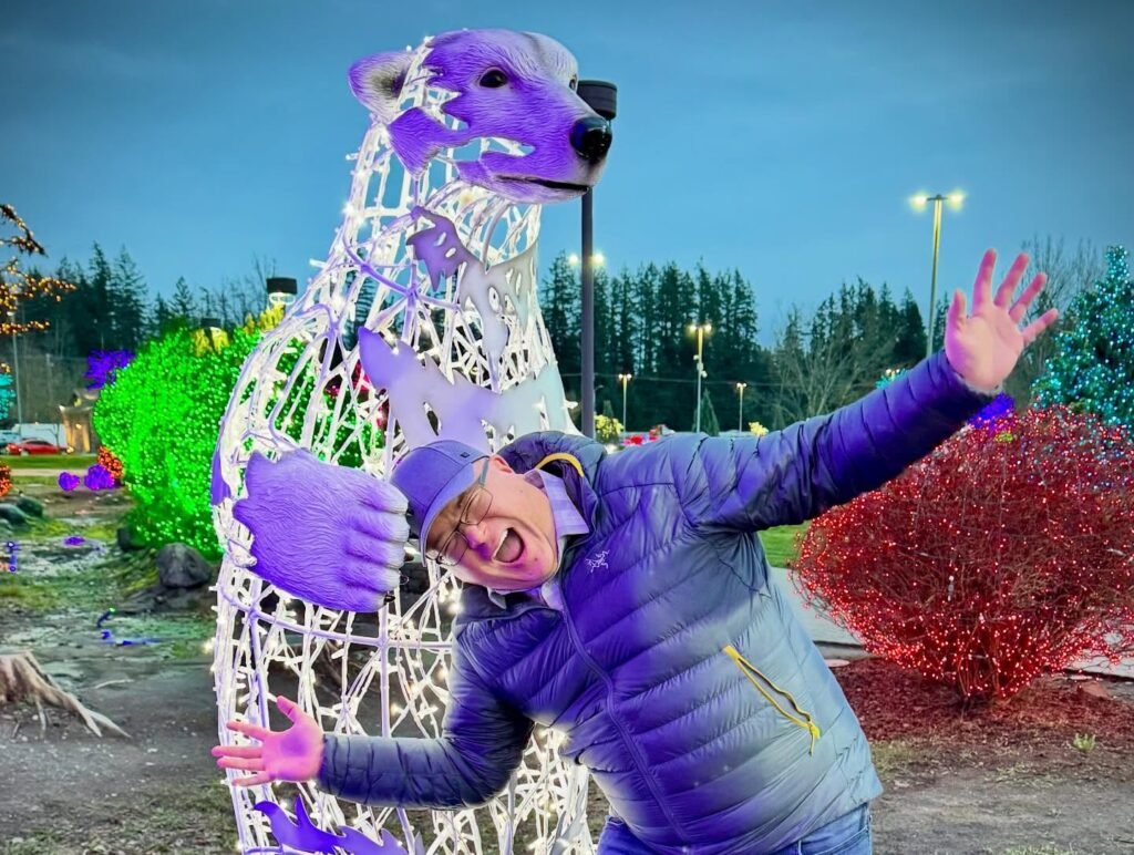 A person wearing a blue jacket poses joyfully with arms outstretched next to a large dog-shaped light sculpture decorated with white, purple, and pink lights.