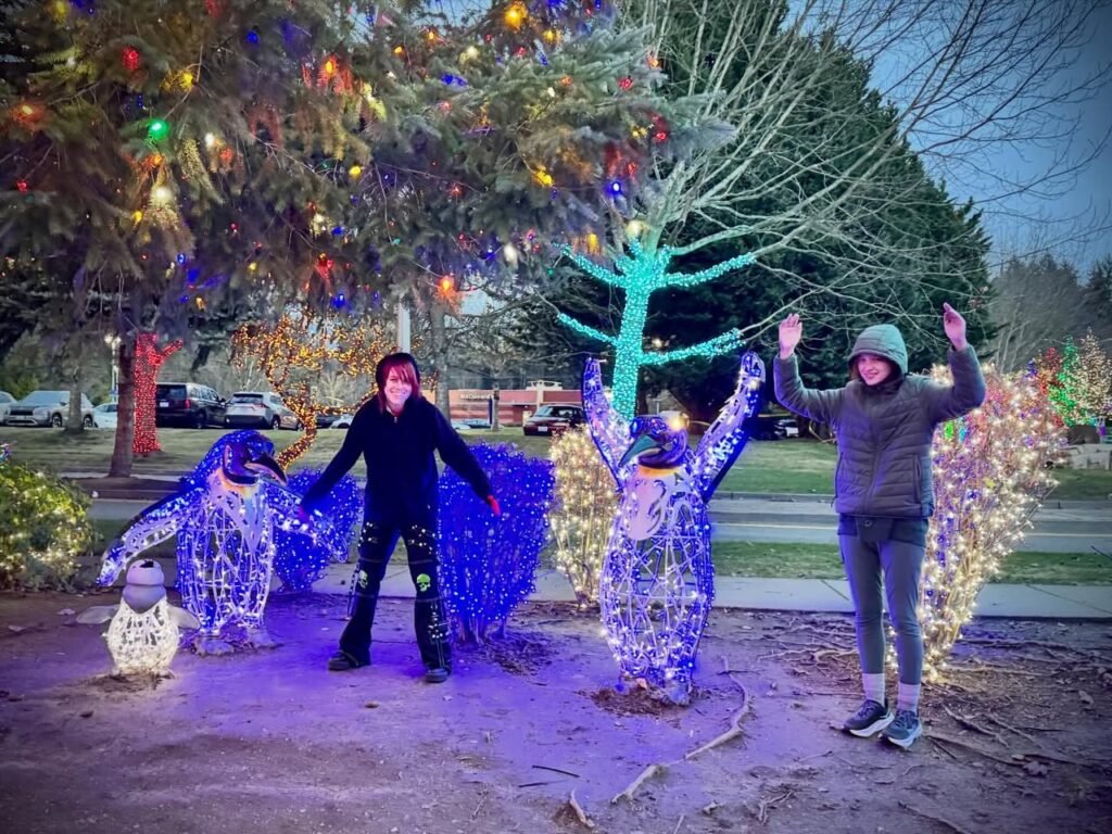 Two people pose beside light-covered penguin and bird sculptures under a Christmas tree at dawn. Vibrant holiday lights highlight the whimsical winter display.