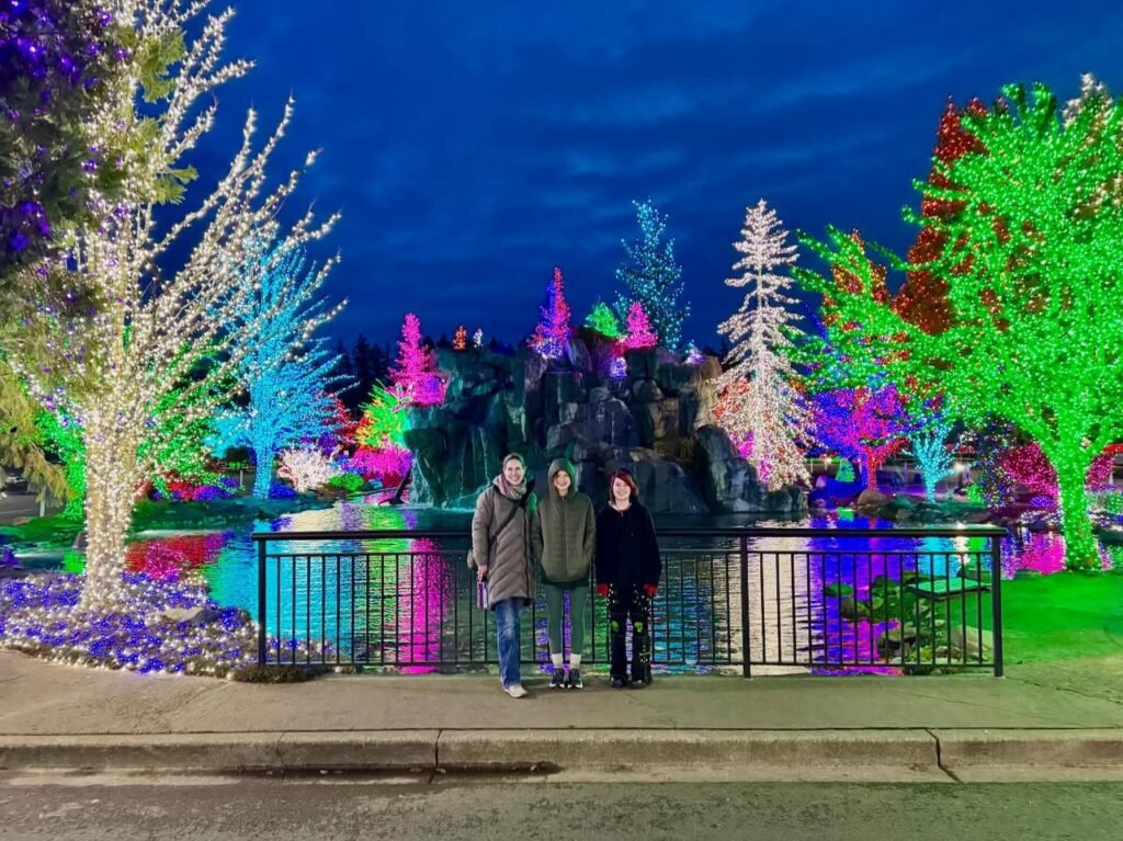 A group of three people stands before a colorful tree light display at dusk.