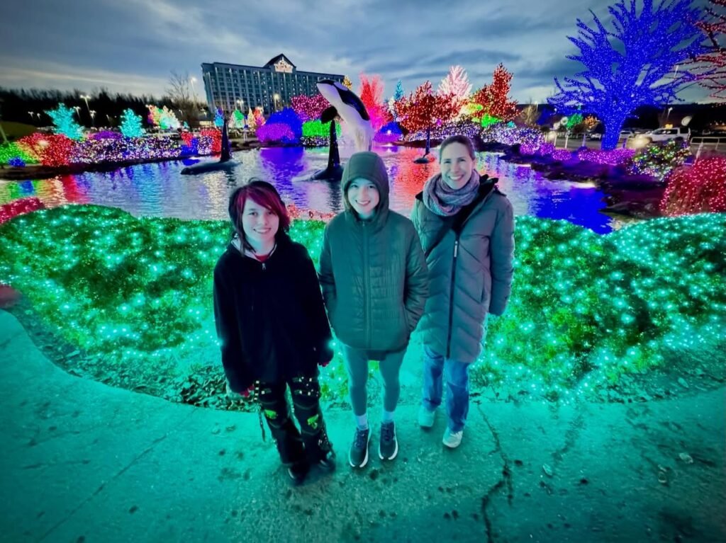 A family enjoys visiting a vibrant holiday light show. The festive atmosphere is highlighted by colorful LEDs and glowing trees.