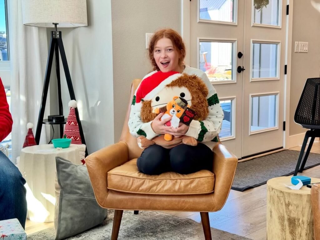 A person wearing a festive vest sits on a leather chair holding a Santa reindeer plushie. The background features a cozy living room with a tripod and green gift boxes.