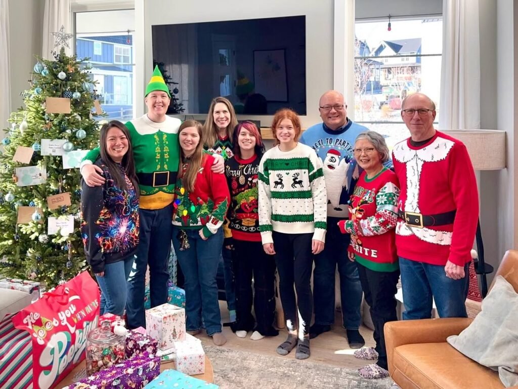 A group of people dressed in festive Christmas sweaters and costumes poses in front of a decorated Christmas tree in a bright living room.