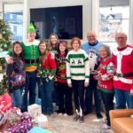 A group of people dressed in festive Christmas sweaters and costumes poses in front of a decorated Christmas tree in a bright living room.