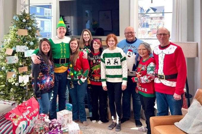 A group of people dressed in festive Christmas sweaters and costumes poses in front of a decorated Christmas tree in a bright living room.