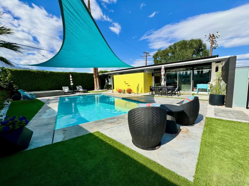 A modern backyard pool area featuring vibrant teal shade sails, wicker furniture, and a yellow accent wall.