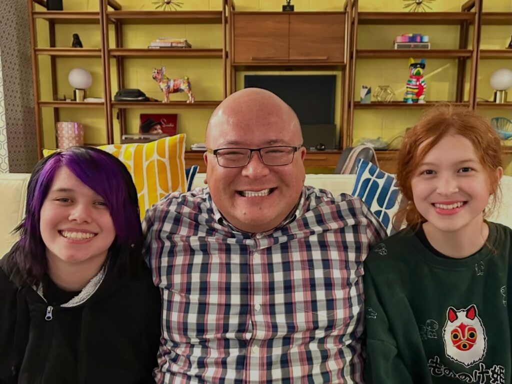 A family of three sits together on a couch in a cozy living room with wooden shelves and personal items in the background.