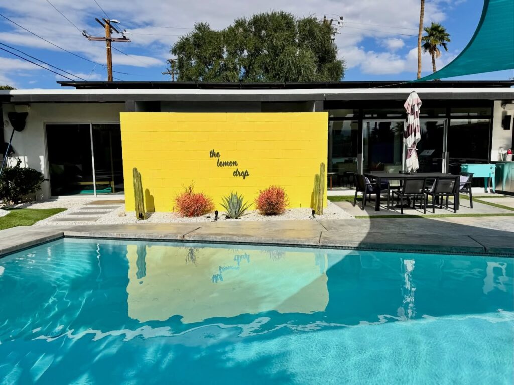 A striking yellow wall features cacti and a modern pool reflects the bright mid-century architecture under a clear blue sky.