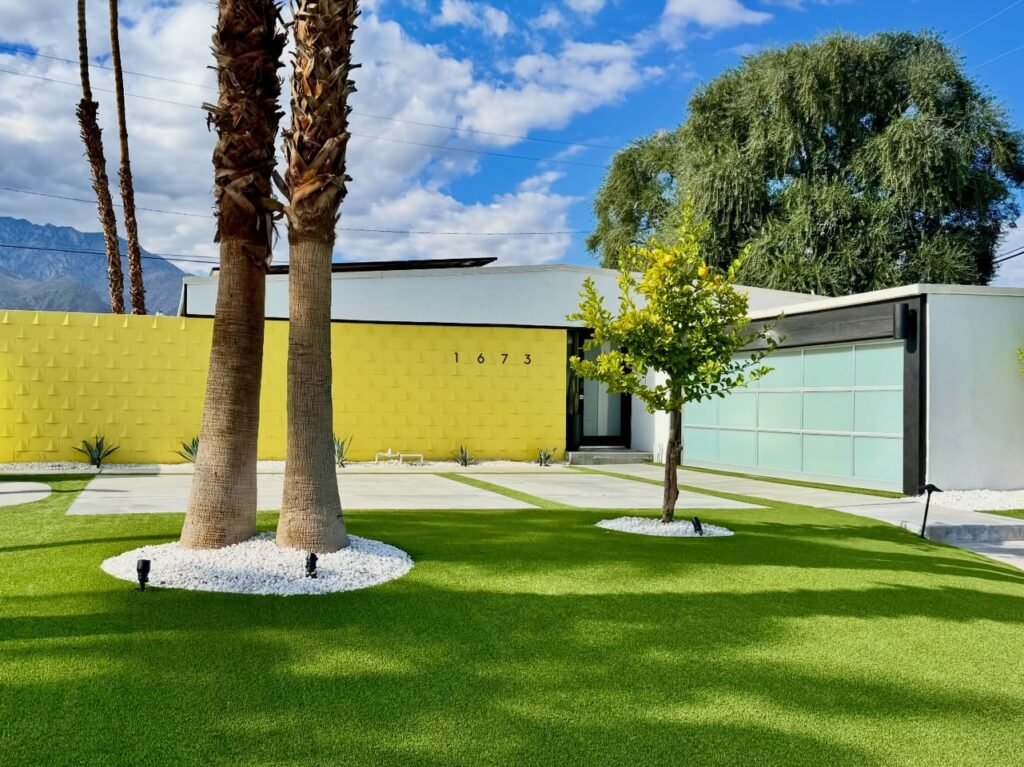 A modern home with a bright yellow wall is set against a backdrop of palm trees and green grass under a sunny blue sky.