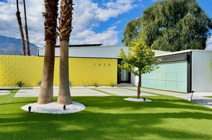 A modern home with a bright yellow wall is set against a backdrop of palm trees and green grass under a sunny blue sky.
