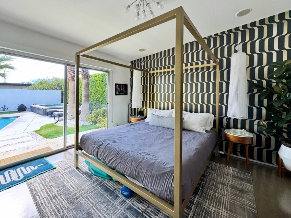 A modern bedroom with a four-poster bed, patterned wallpaper, and glass doors opening to a pool area.