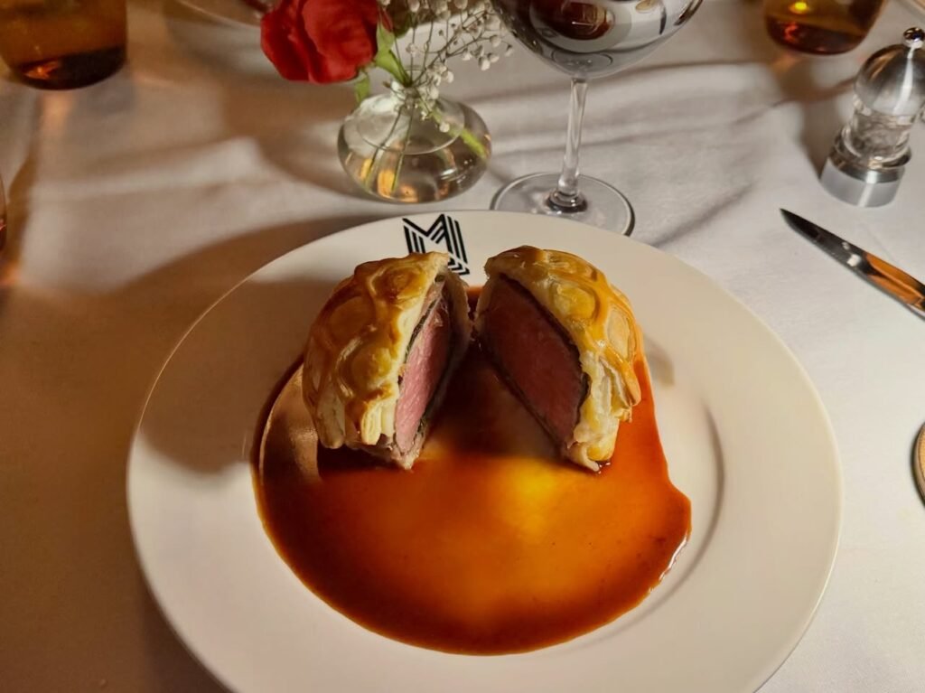 Beef Wellington is served on a white plate with a rich red sauce. A table setting with wine glasses and flowers is visible.