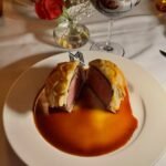 Beef Wellington is served on a white plate with a rich red sauce. A table setting with wine glasses and flowers is visible.