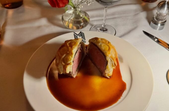 Beef Wellington is served on a white plate with a rich red sauce. A table setting with wine glasses and flowers is visible.
