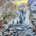 Birthday Dinner at Mr. Lyons A scenic view of a waterfall cascading down a rocky cliff with hikers at its base.