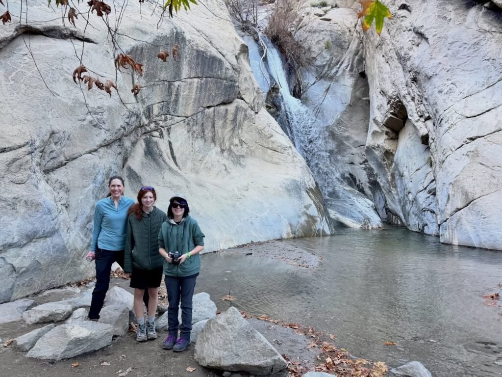 A family of three poses in front of a waterfall in a natural granite canyon. The rocky terrain and shallow stream create a beautiful backdrop for this outdoor adventure.