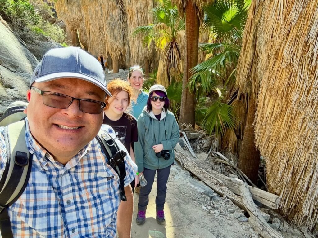 A group of hikers enjoys a trail lined with palm trees and lush vegetation. One hiker takes a selfie from the front of the line.