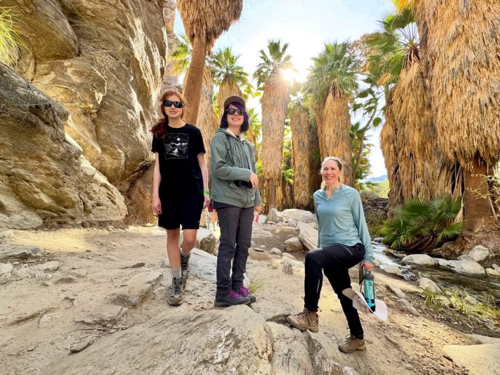 A group of three people posing among tall palm trees and rocky canyon walls on a sunny day.