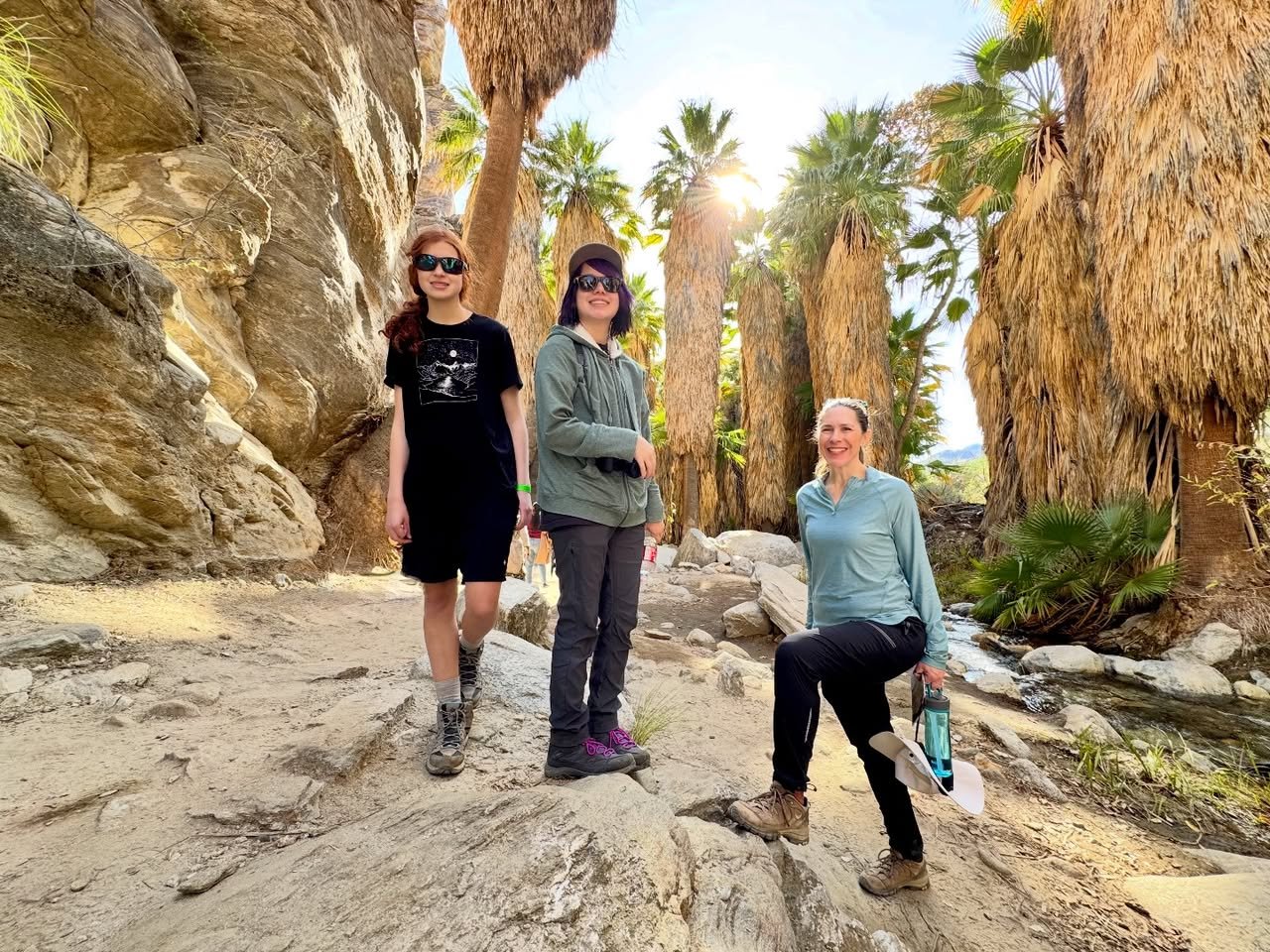 Andreas Canyon A group of three people posing among tall palm trees and rocky canyon walls on a sunny day.