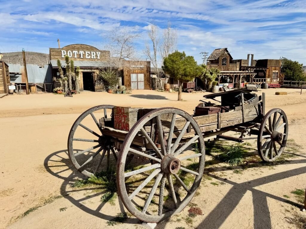 A rustic wooden wagon sits in a dusty street in front of the antique western building labeled Pottery.