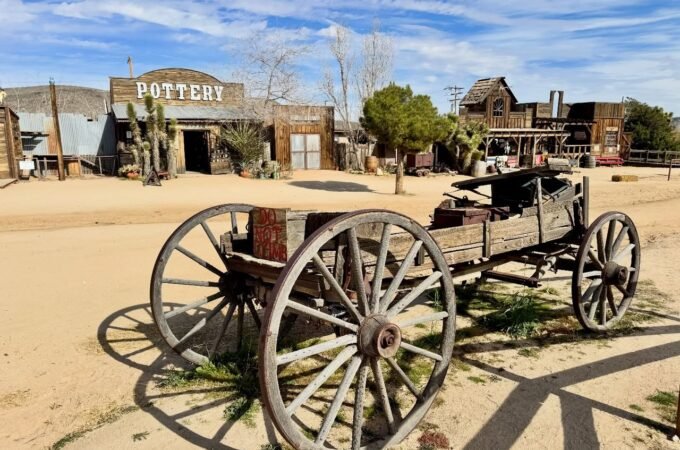 A rustic wooden wagon sits in a dusty street in front of the antique western building labeled Pottery.