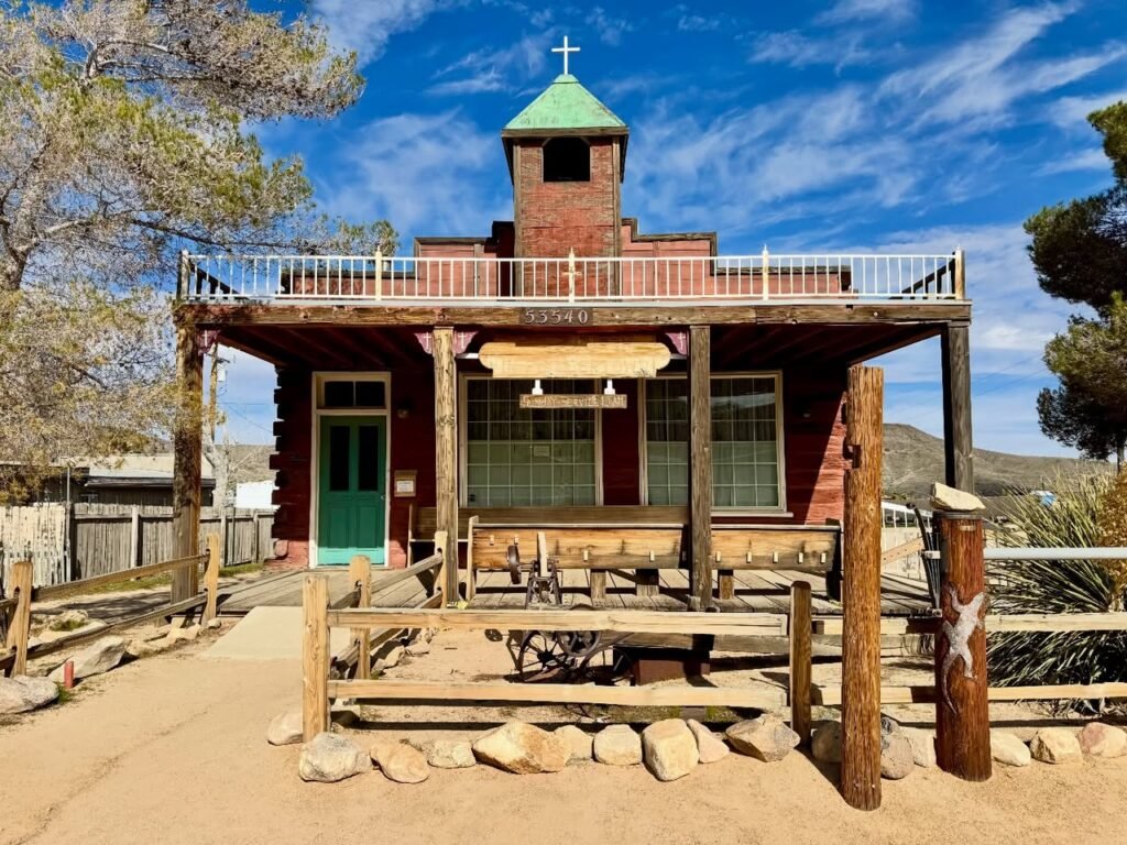 A weathered wooden railroad station with a distinct bell tower topped by a cross, set against a blue sky.