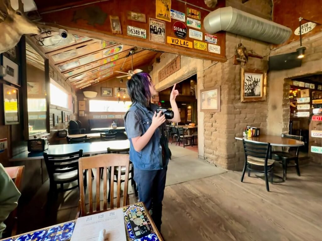 A person holding a camera looks up at license plates and decorations hanging from the ceiling of a rustic restaurant.
