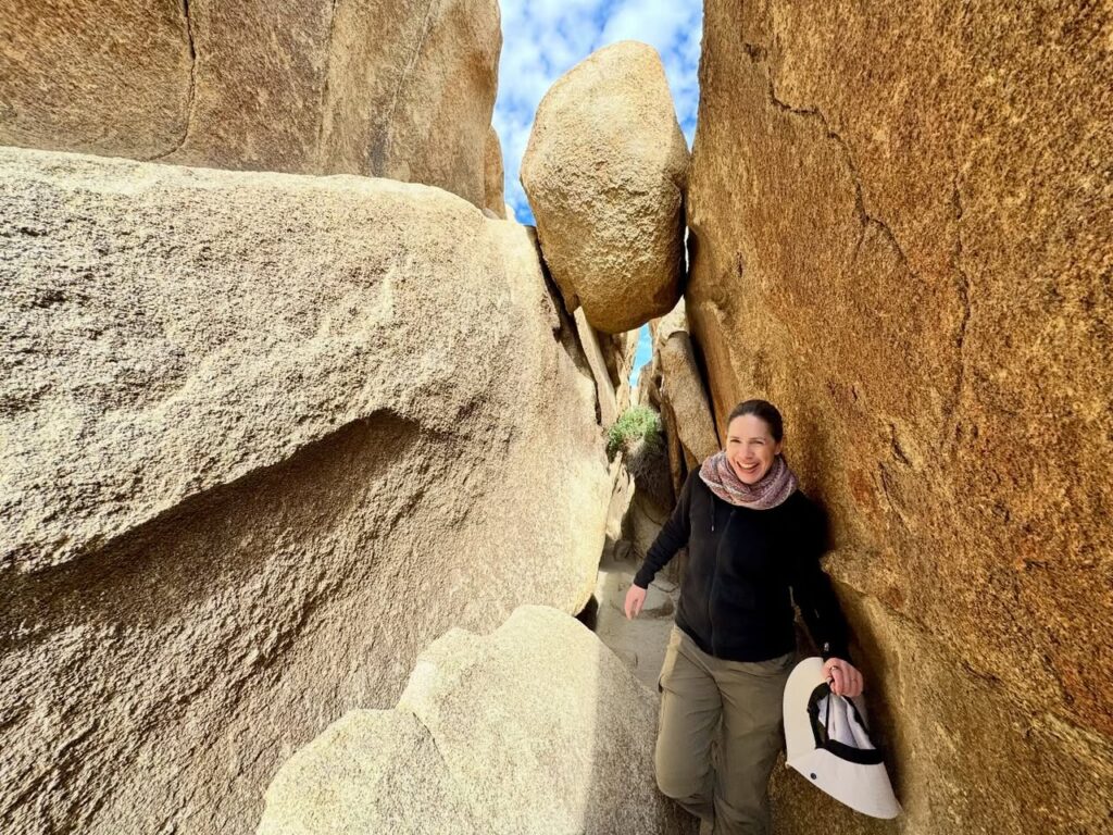 A person stands in a narrow rocky passage between massive granite boulders, exploring a natural outdoor landscape.