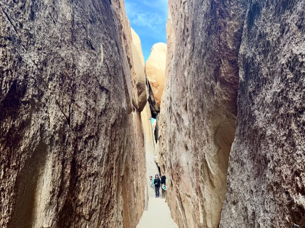 Walk through a narrow rock passage surrounded by towering weathered granite walls under a clear blue sky.