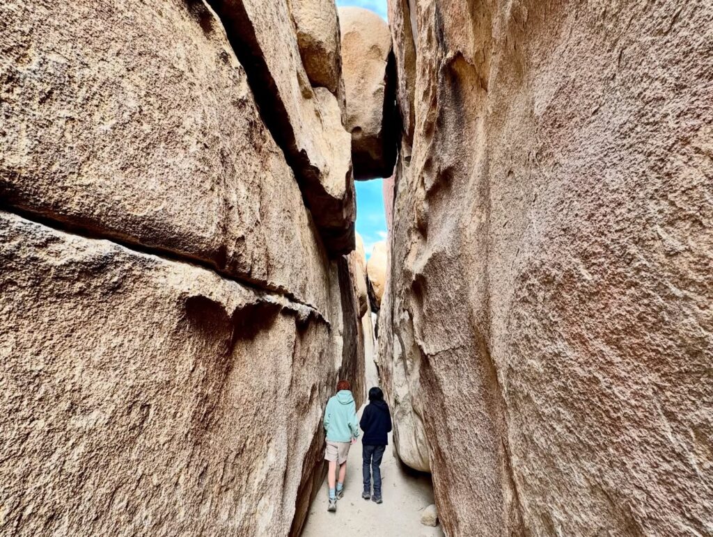 A hiker walks through a narrow slot canyon with towering rock walls framing the path.