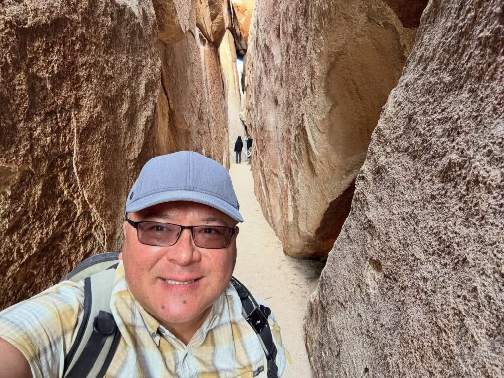 A traveler wearing a blue cap and glasses captures a selfie while hiking a narrow slot canyon. The rugged rock formations and sandy path highlight the natural environment.