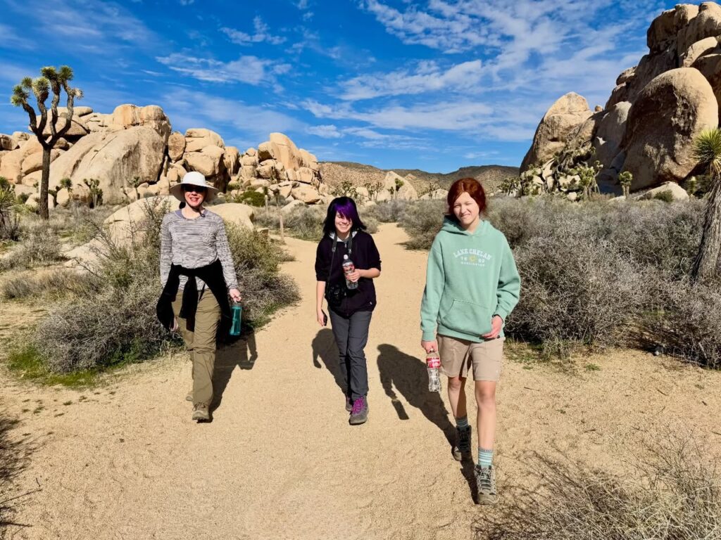 Three people walking on a sandy desert path through a landscape of boulders and Joshua trees under a blue sky.