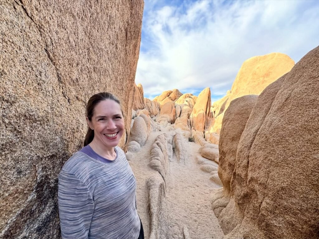 A woman smiles while hiking through a narrow canyon of smooth granite boulders under a sunny sky. The image showcases unique rock formations and outdoor adventure in the desert.