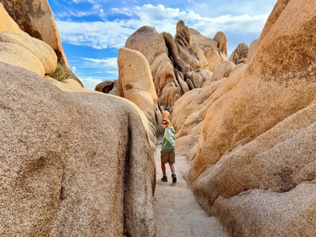 A hiker stands in a narrow pass between large smooth granite boulders under a blue sky with clouds.