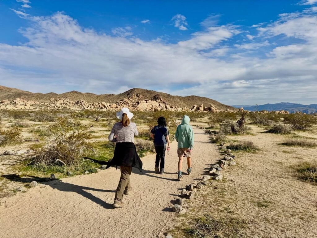 Three hikers walking along a dirt trail in a desert landscape with hills and scattered clouds overhead. This scene captures the essence of outdoor travel and exploration.