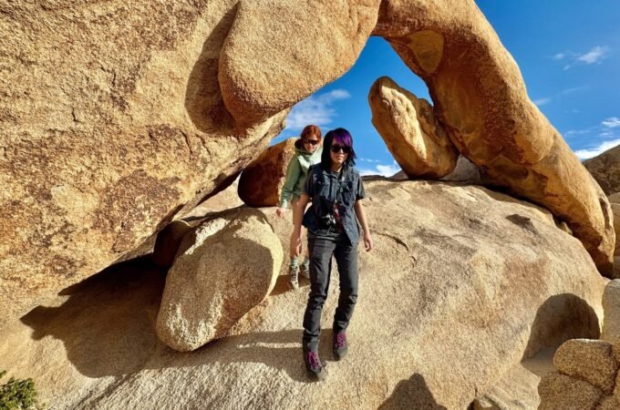 Two hikers pose beneath a massive natural rock arch on a sunny day.