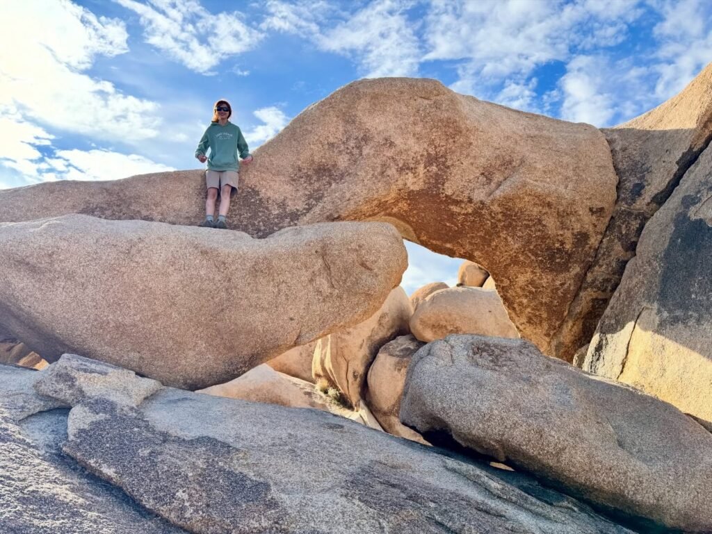 A person standing on massive granite boulders in a desert landscape. Wide panoramic view capturing the scale of the natural rock formation under a blue sky.