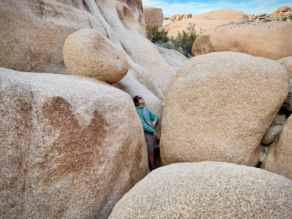 A person stands among large, smooth granite boulders in a desert landscape. Sparse vegetation grows between the rocks under a blue sky.