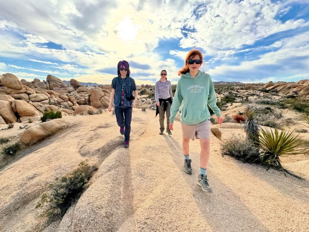 Three hikers walking along a sandy trail in a desert landscape. The scene features large granite boulders and a wide sky with scattered clouds.