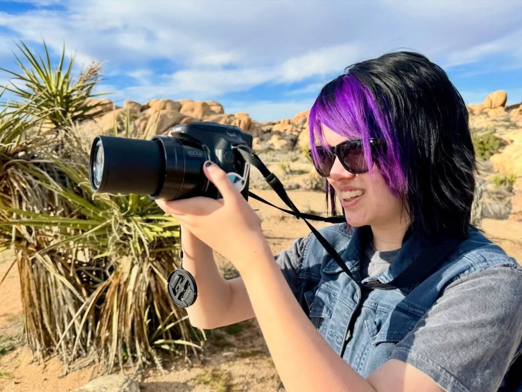 A photographer with vibrant purple and black hair uses a DSLR camera in a rocky desert landscape.