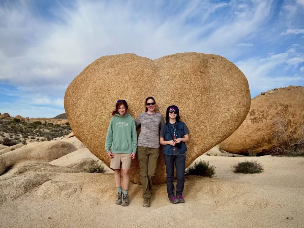 A group of three travelers posing in front of a massive heart-shaped boulder in a desert landscape.